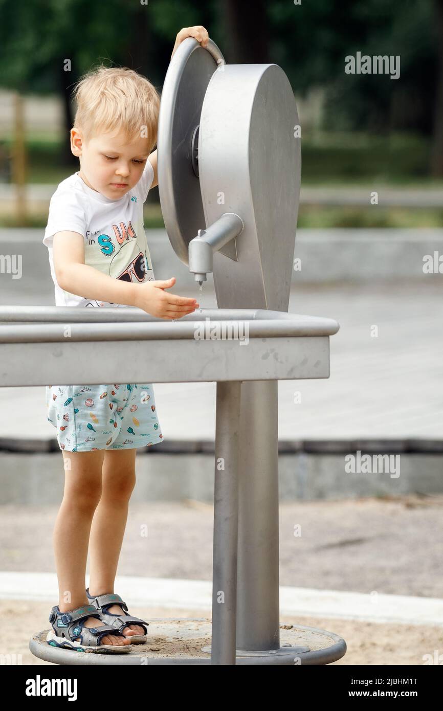 A happy little boy plays with a water tap in a city park. Special water equipment for children's games on a hot summer day outdoors. Vertical photo. Stock Photo