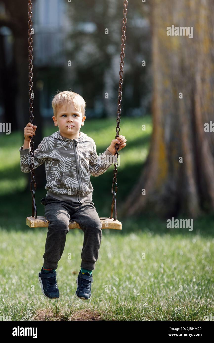 A sad, dreaming little boy swings in a city park under a big tree. Vertical photo. Stock Photo