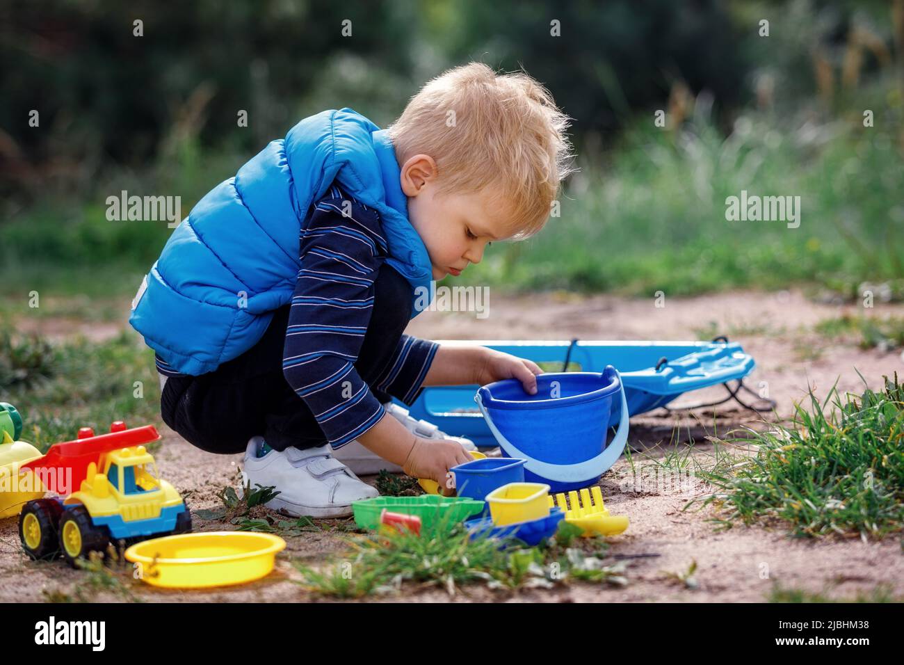 Child squat on the ground playing with sand toys. The child digs the sand into a plastic bucket. Summer outdoor activity for kids. Leisure Time. Stock Photo