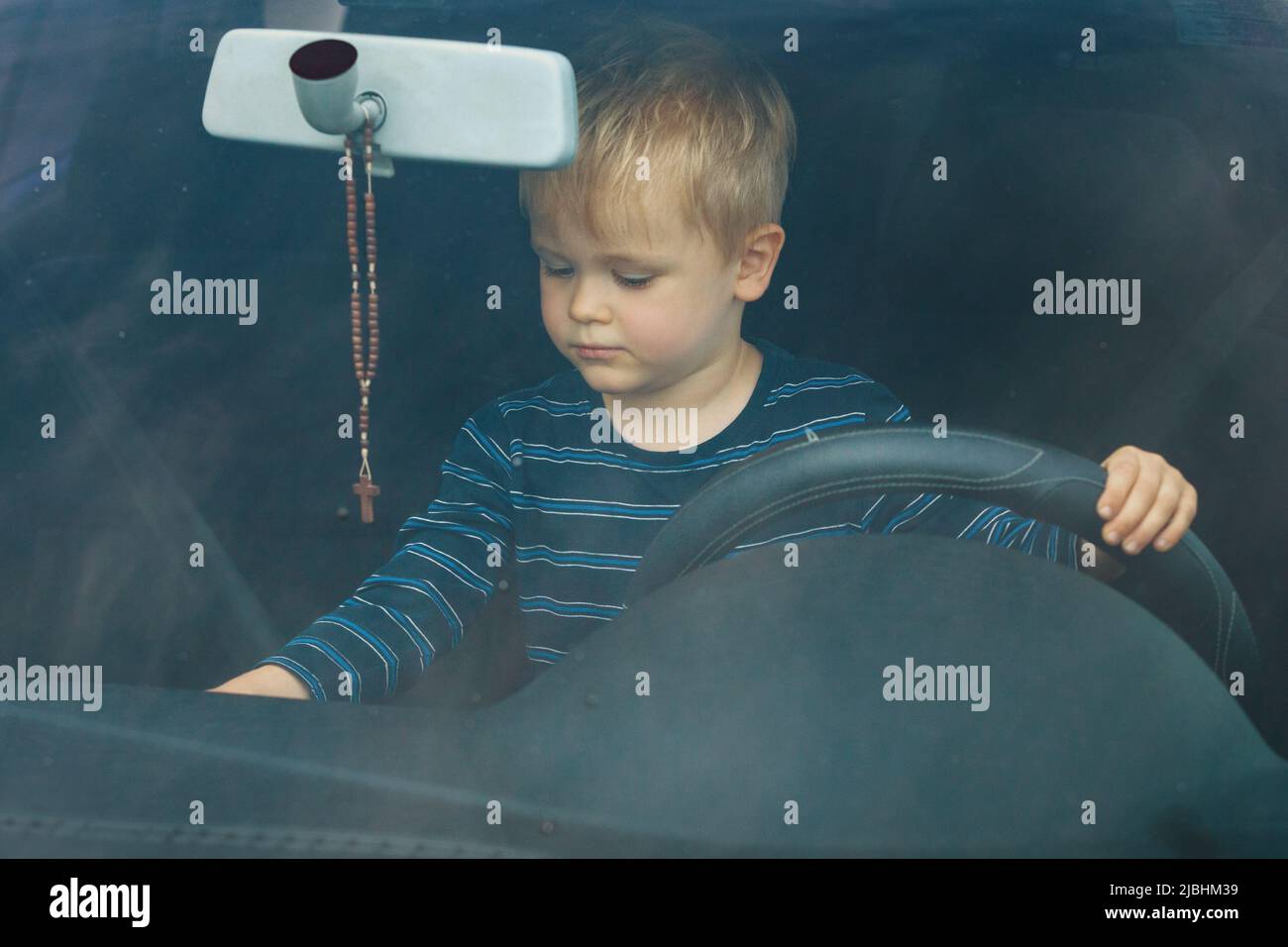 Cute little boy driving fathers car. Portrait of a child sitting in a car behind the wheel from the front. The boy plays and imagines he is driving, h Stock Photo