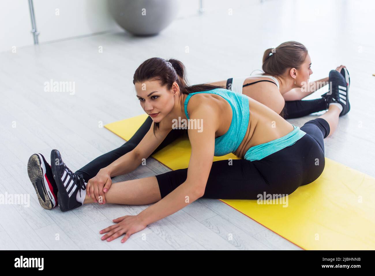 Two slim girls taking part in yoga class working in pair doing partner side seated wide angle pose called parsva upavistha konasana in sports center. Stock Photo