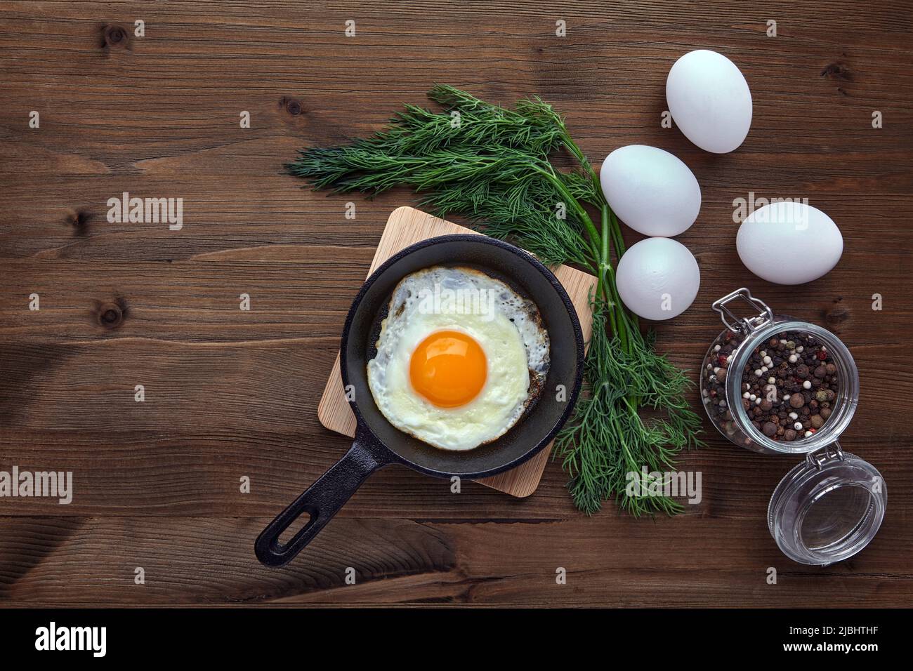 Fried egg in a small skillet with a stand, a bunch of dill, a jar of  peppercorns and whole white eggs on a brown wooden table Stock Photo - Alamy
