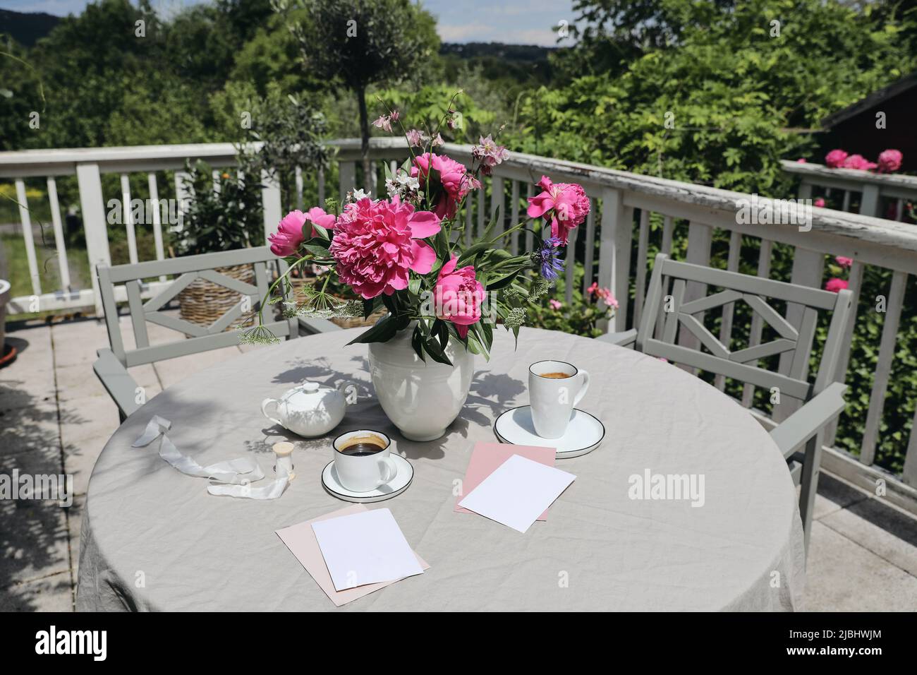 Summer breakfast on terrace. Cups of coffeee, blank greeting cards and peonies bouquet. Gey wooden table. Outdoor dining. Blurred green garden, yard Stock Photo