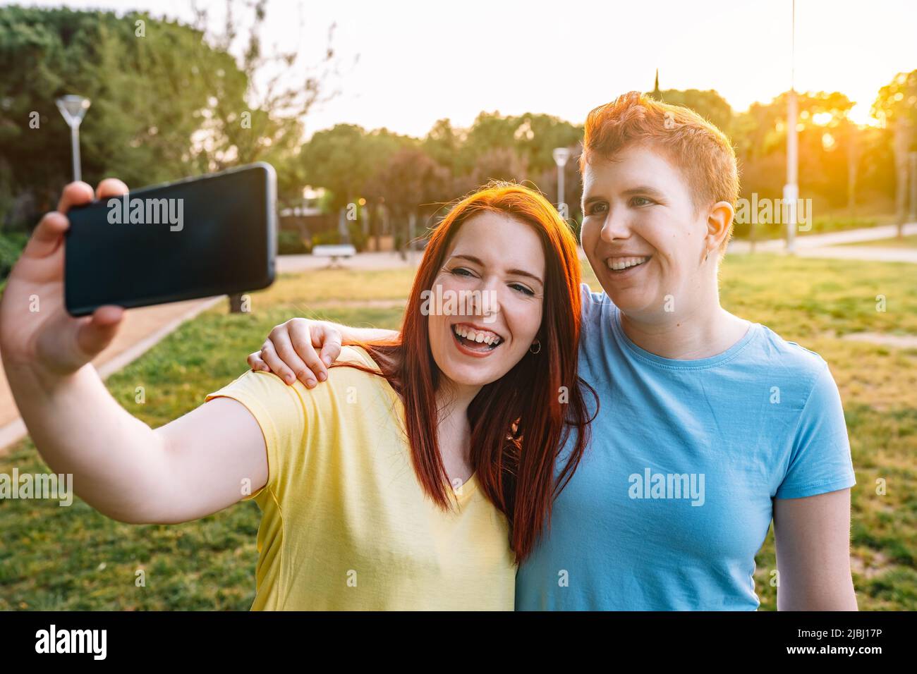 couple of friends taking a selfie with their smartphones, laughing out loud in a public park in the city at sunset. couple of young girls enjoying the Stock Photo