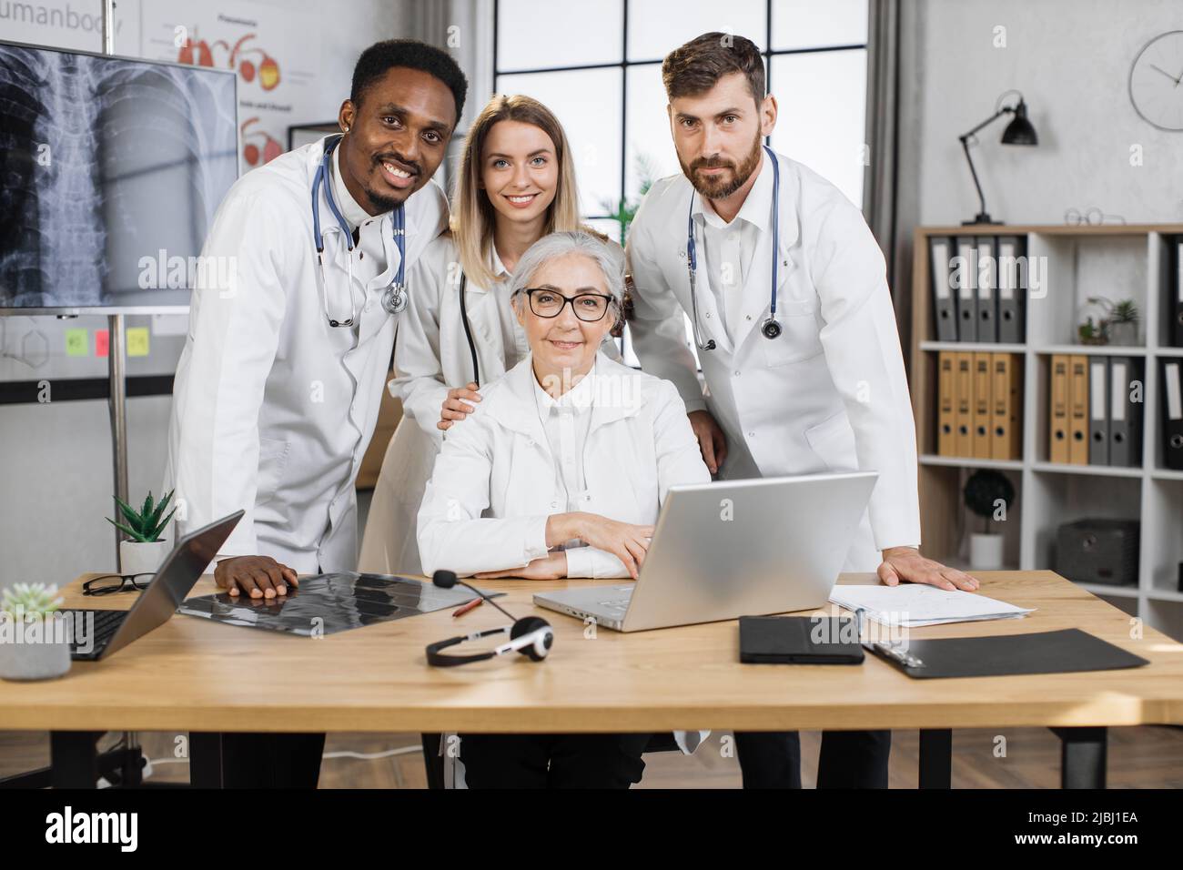 Happy researchers posing on camera with smile in front of laptop while working on new scientific project. Multiethnic lab staff making medical discoveries in modern pharmaceutical company. Stock Photo