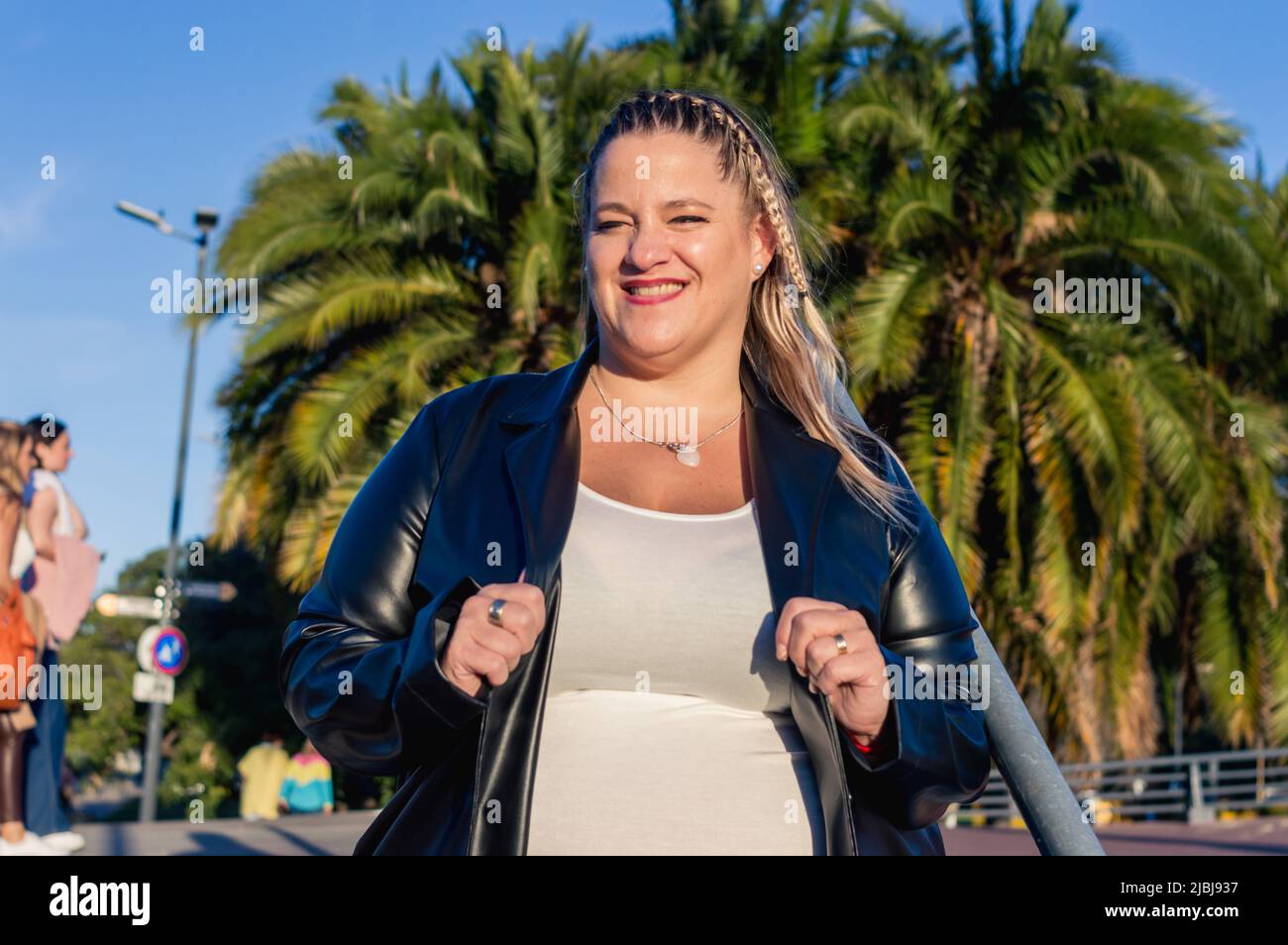 portrait argentinian blonde adult latin woman, standing outdoors holding jacket lapel with hands, looking at camera smiling, plus size model, lifestyl Stock Photo
