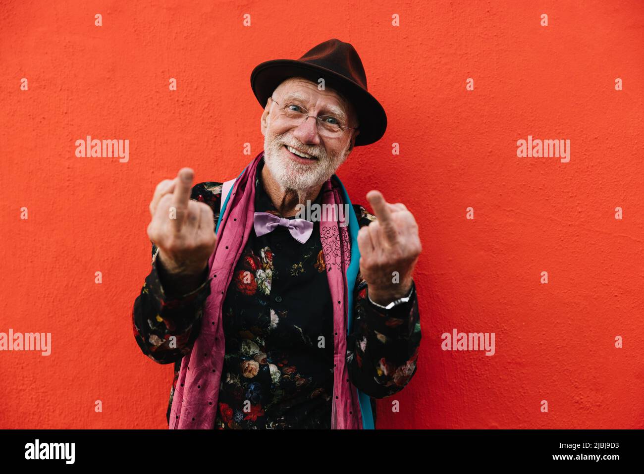 Carefree senior man showing his middle fingers while standing against a red background. Mature man looking at the camera while making a swear gesture. Stock Photo