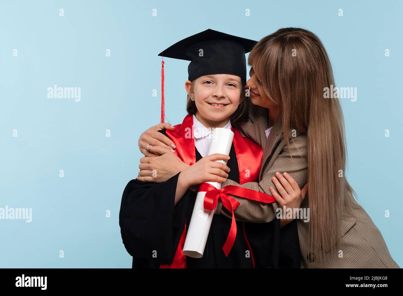 Little girl graduate celebrating graduation. Child wearing graduation cap and ceremony robe Holding Certificate. Mom hugs and congratulations daughter Stock Photo
