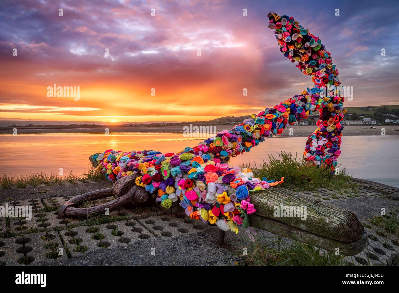 Appledore, North Devon, England. Tuesday 7th June 2022. The sun rises behind the landmark anchor on the quay in Appledore. The anchor has been decorated with hundreds of colourful crocheted flowers by the residents of the North Devon coastal village. Credit: Terry Mathews/Alamy Live News Stock Photo