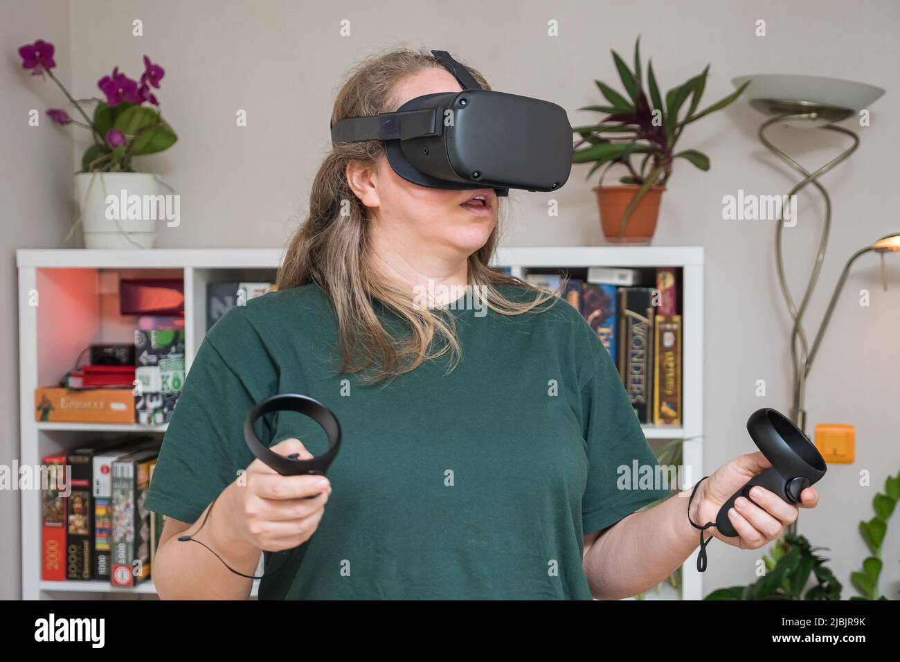 A girl in a VR helmet wearing a green T-shirt and controllers in her hands travels through metacenter. In background are shelves with board games, flo Stock Photo