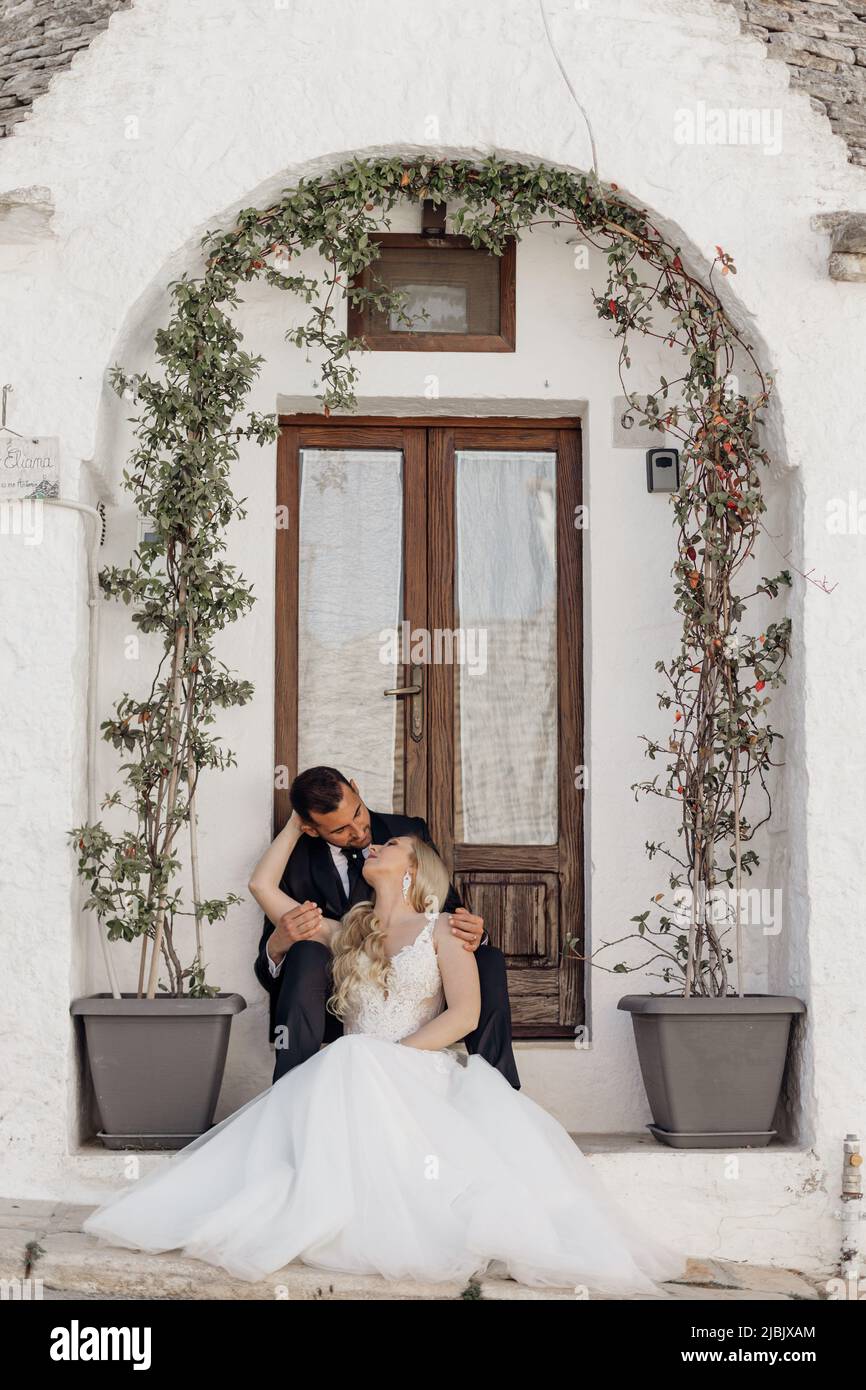 Portrait of blonde bride in white dress and brunet groom in suit kissing and sitting near old city house with arch from plants in flowerpots in Italy Stock Photo