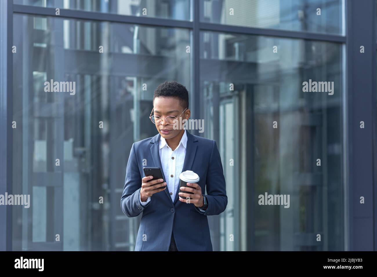 A confident African-American lady walks around the business district and uses the phone. Holding a cup of coffee, dressed in formal clothes. Attractiv Stock Photo