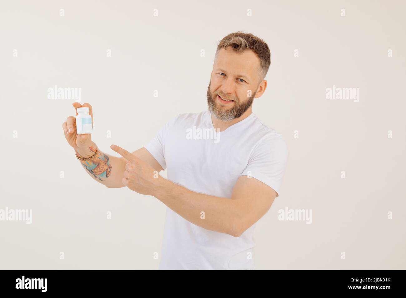 Portrait of young smiling man advertising useful vitamins for men, white background. Holding in hand and pointing at jar of pills, disease treatment Stock Photo