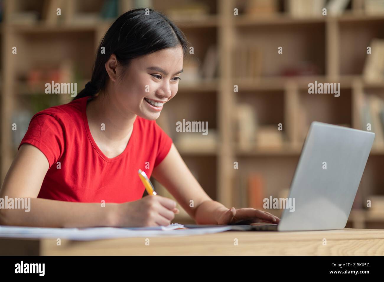 Smiling happy teenage japanese girl doing homework at table with laptop in room interior Stock Photo
