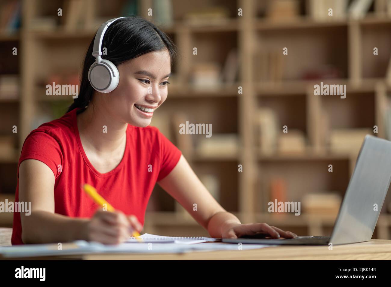 Cheerful teen korean female in headphones listen lesson at laptop in room interior, profile