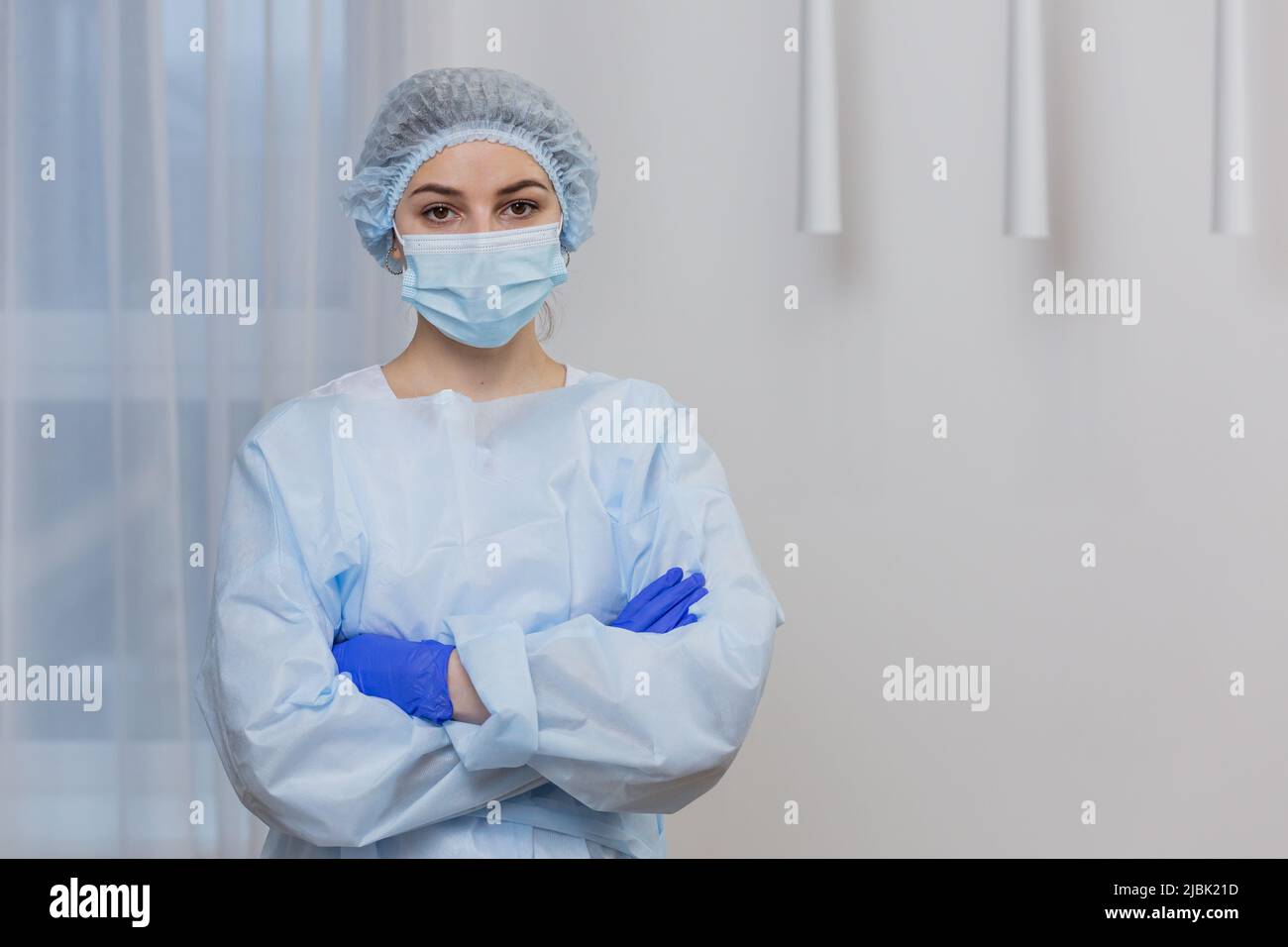 Portrait of a young female doctor in a mask, surgical gown and hat, standing with arms crossed, looking at camera Stock Photo