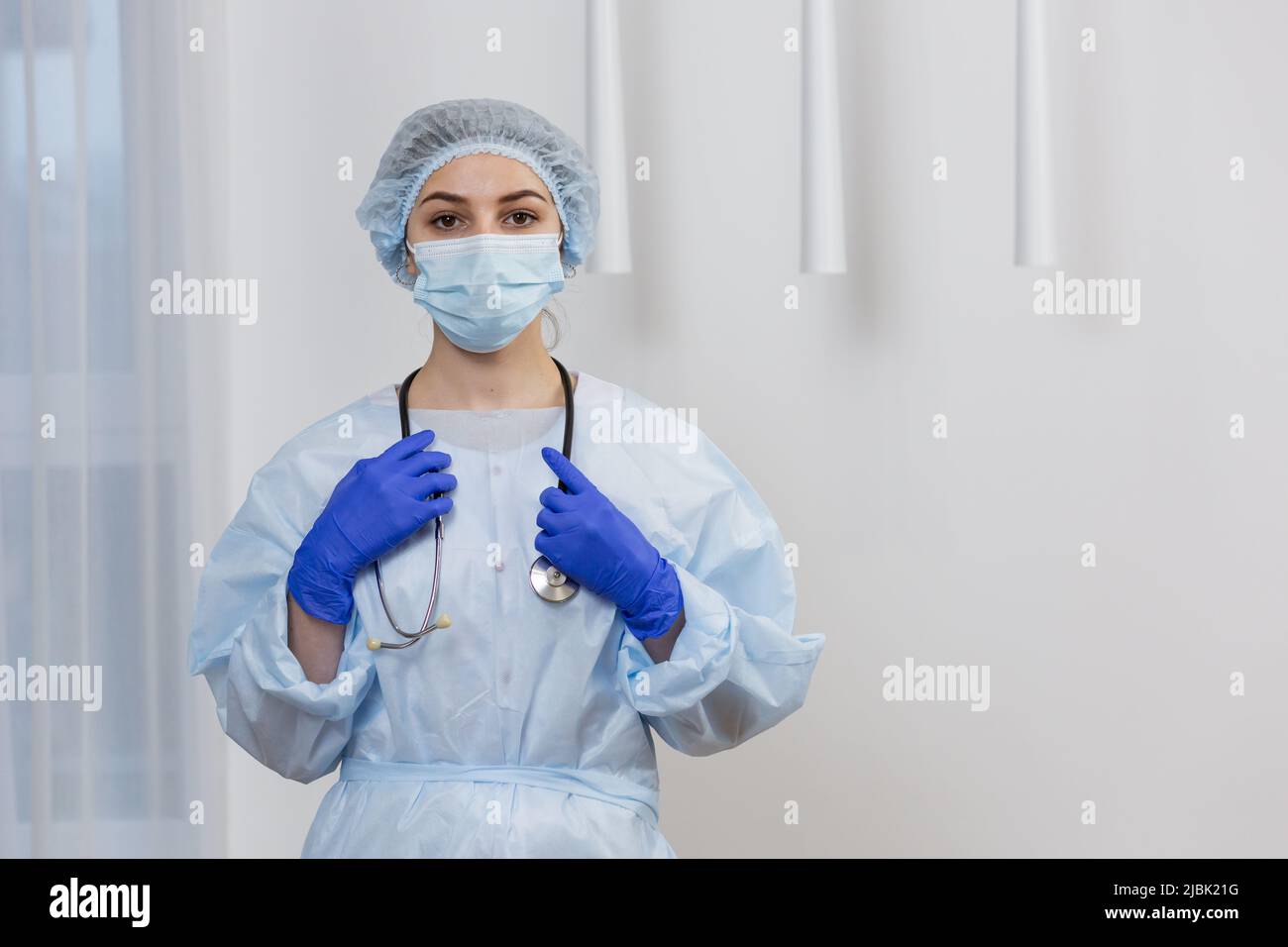 Portrait of young female doctor in mask, surgical gown and hat, standing holding stethoscope, looking at camera Stock Photo