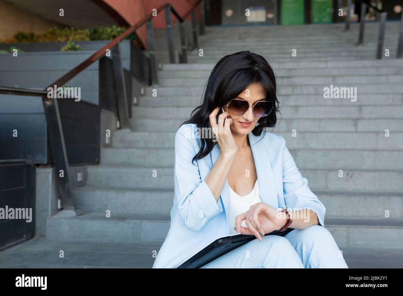beautiful young business woman sitting on the stairs on the street near the office center, talking on the phone, making a video call, waiting for some Stock Photo