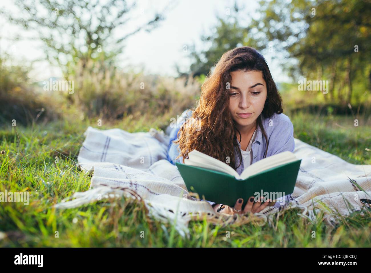 Beautiful young student girl reads a book, studies, lying on the grass in nature Stock Photo