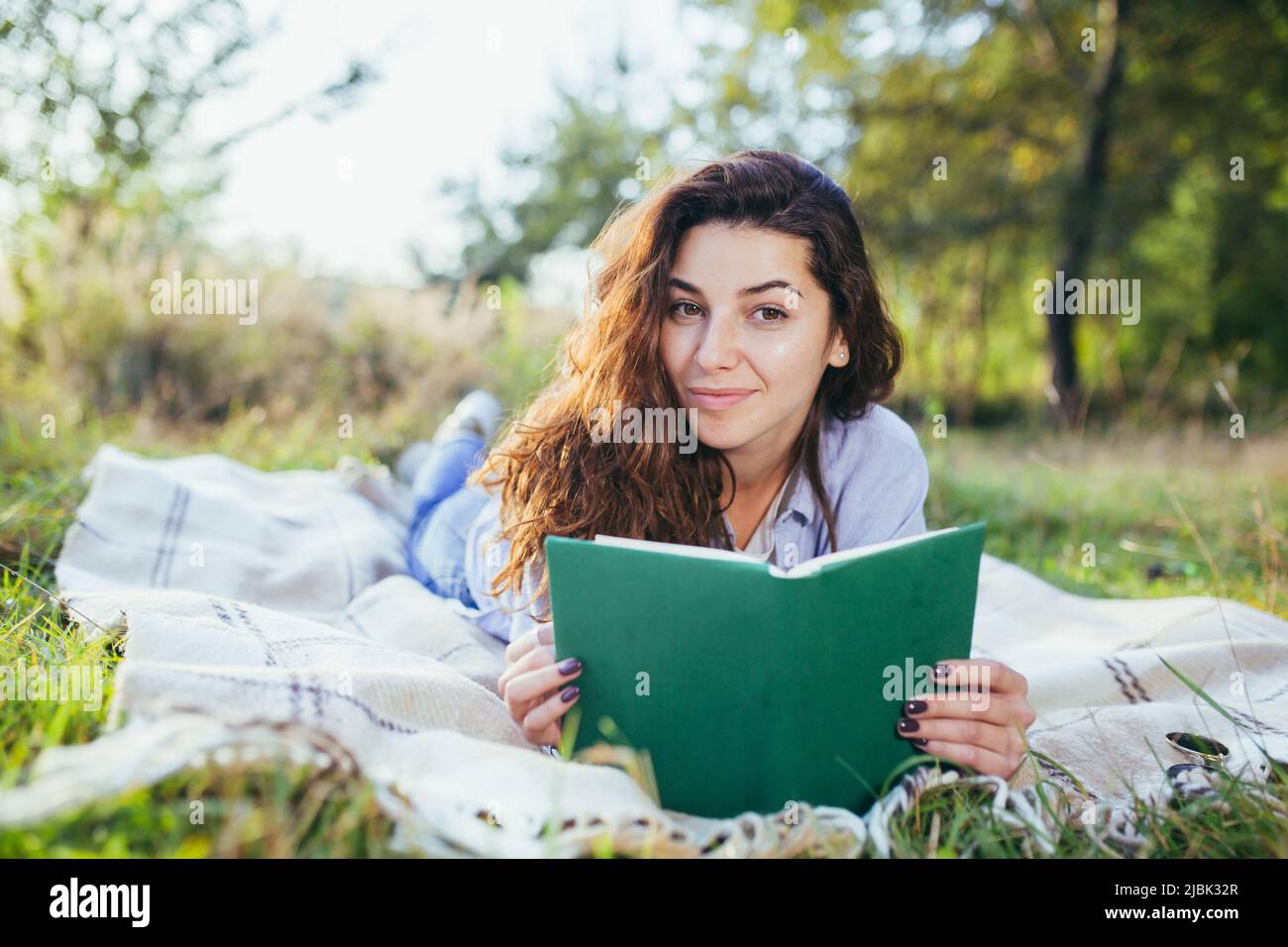 Beautiful young student girl reads a book, studies, lying on the grass in nature Stock Photo