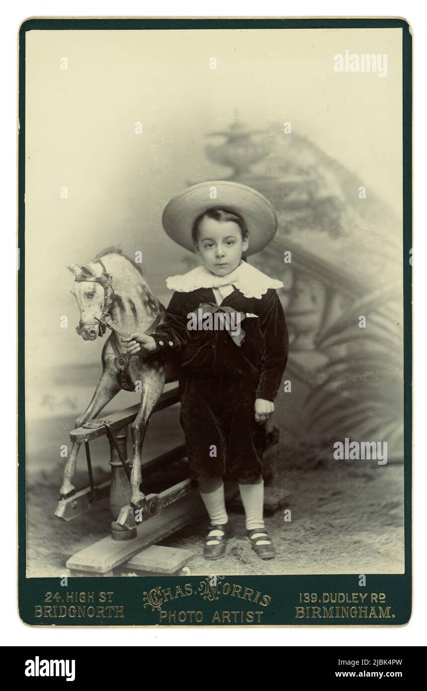 Original charming Victorian era Cabinet card of a cute young boy wearing a velvet suit with a lace collar (Little Lord Fauntleroy style) standing next to a beautiful rocking horse.  From the studio of Chas (Charles) Norris, with studios at  High St. Bridgnorth, Shropshire and Dudley Rd, Birmingham, U.K. Circa late 1890's, 1900. Stock Photo