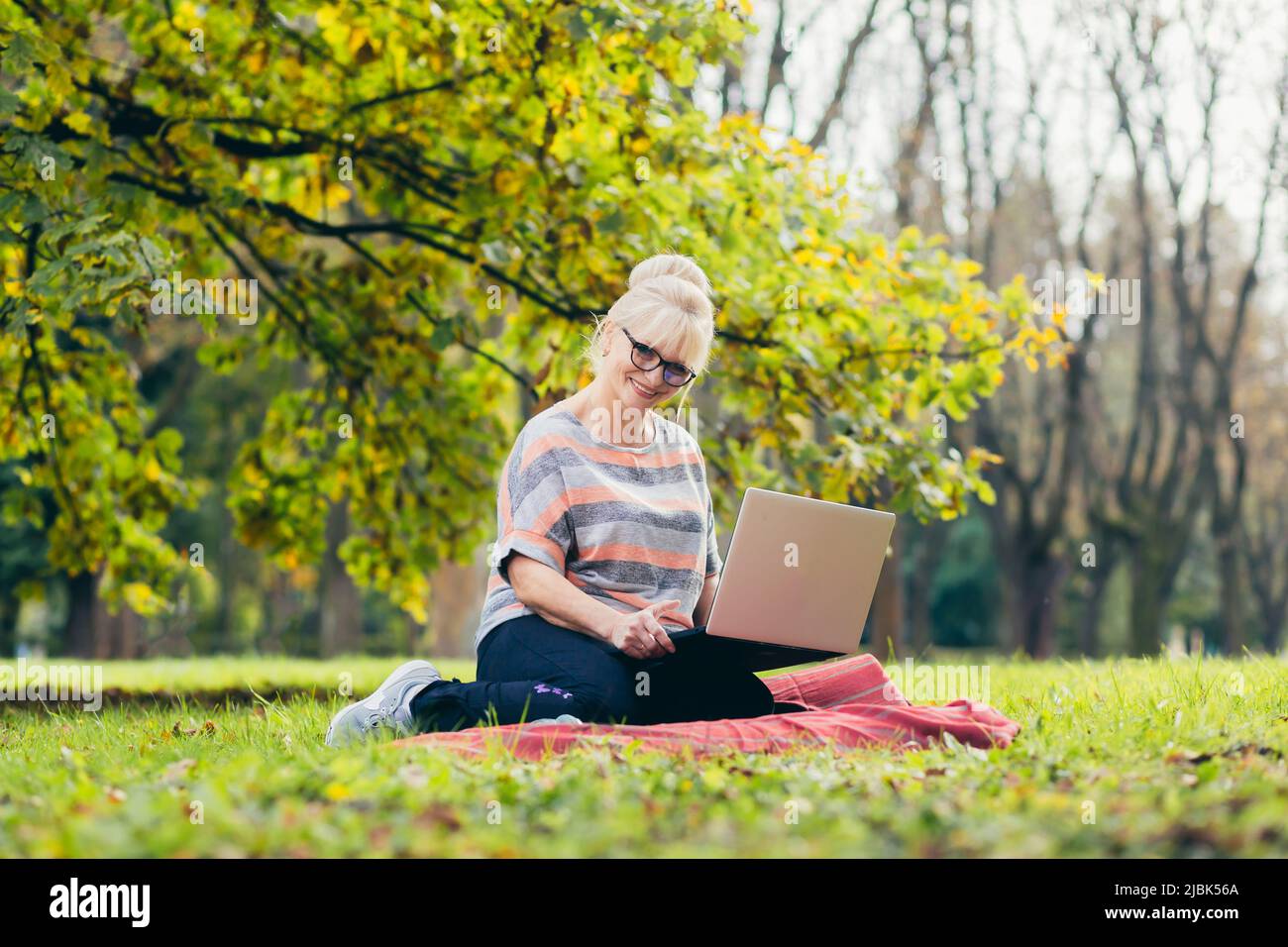 Beautiful blonde woman, senior, sitting on the grass in the park uses a laptop, works, studies, talks on video link Stock Photo