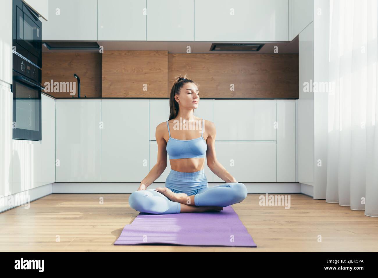Yoga time. Portrait of a young beautiful woman practicing yoga at home, sitting on a mat in the lotus position, looking at the camera, smiling, relaxe Stock Photo