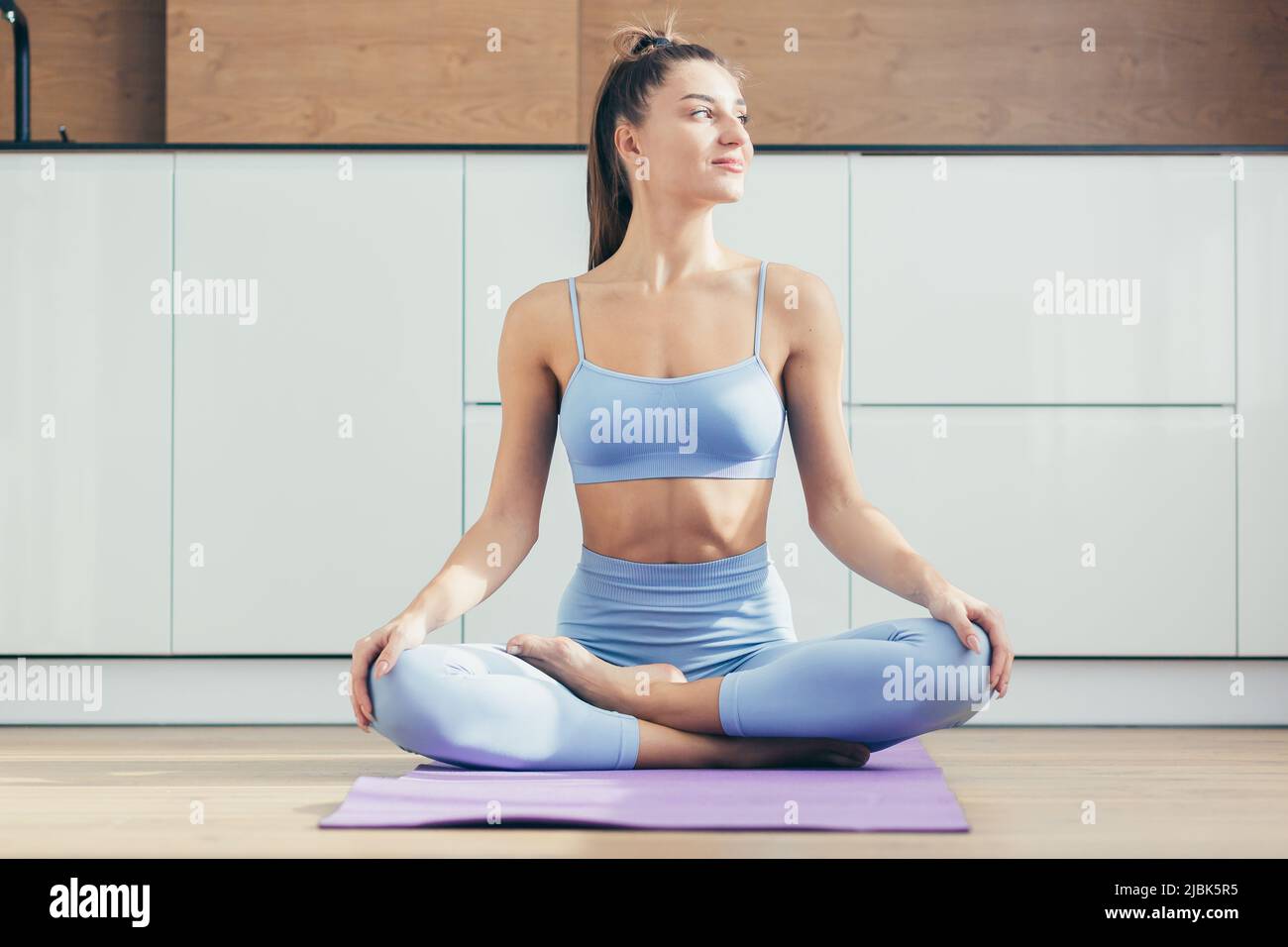 Yoga time. Portrait of a young beautiful woman practicing yoga at home, sitting on a mat in the lotus position, looking at the camera, smiling, relaxe Stock Photo