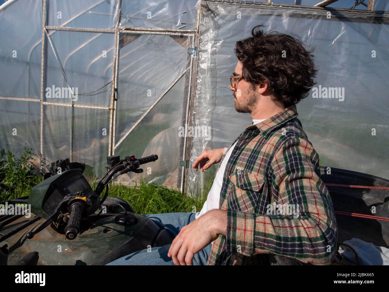 Young caucasion male farmer on an off road farm vehicle contemplating beside an organic vegetable garden greenhouse in natural golden hour sunlight wi Stock Photo