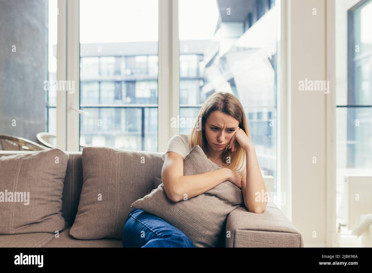 Upset young woman sitting on sofa at home, hugging pillow, feeling bad