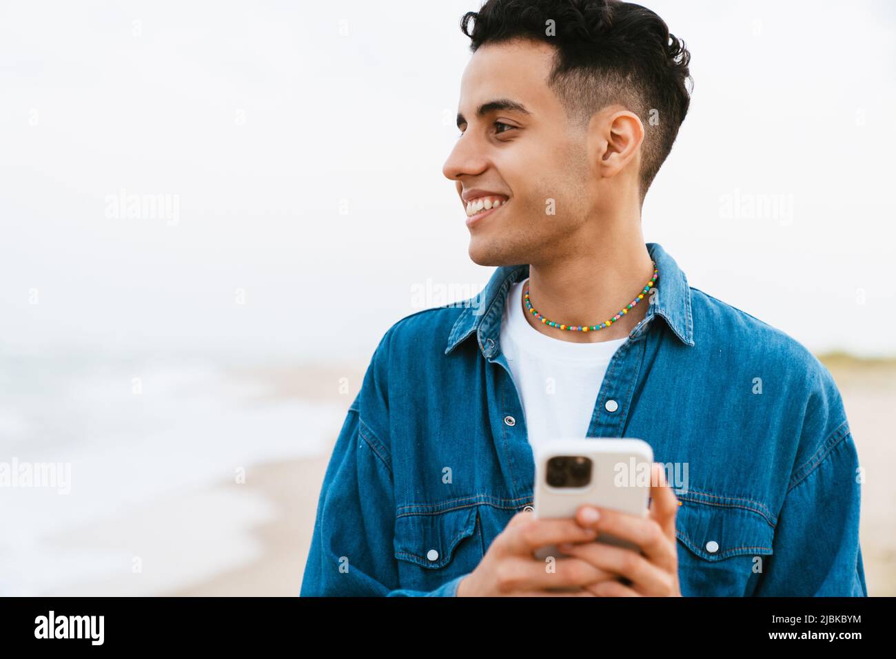 Young middle eastern man smiling while using cellphone at summer beach Stock Photo