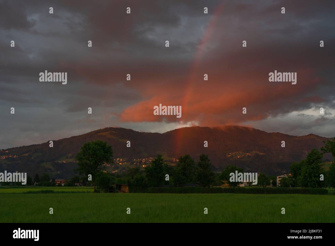 leuchtendes Abendrot mit Regenbogen nach dem Sonnenuntergang in Dornbirn, Vorarlberg, Austria. rote und graublaue Wolken über den grünen Wiesen Stock Photo