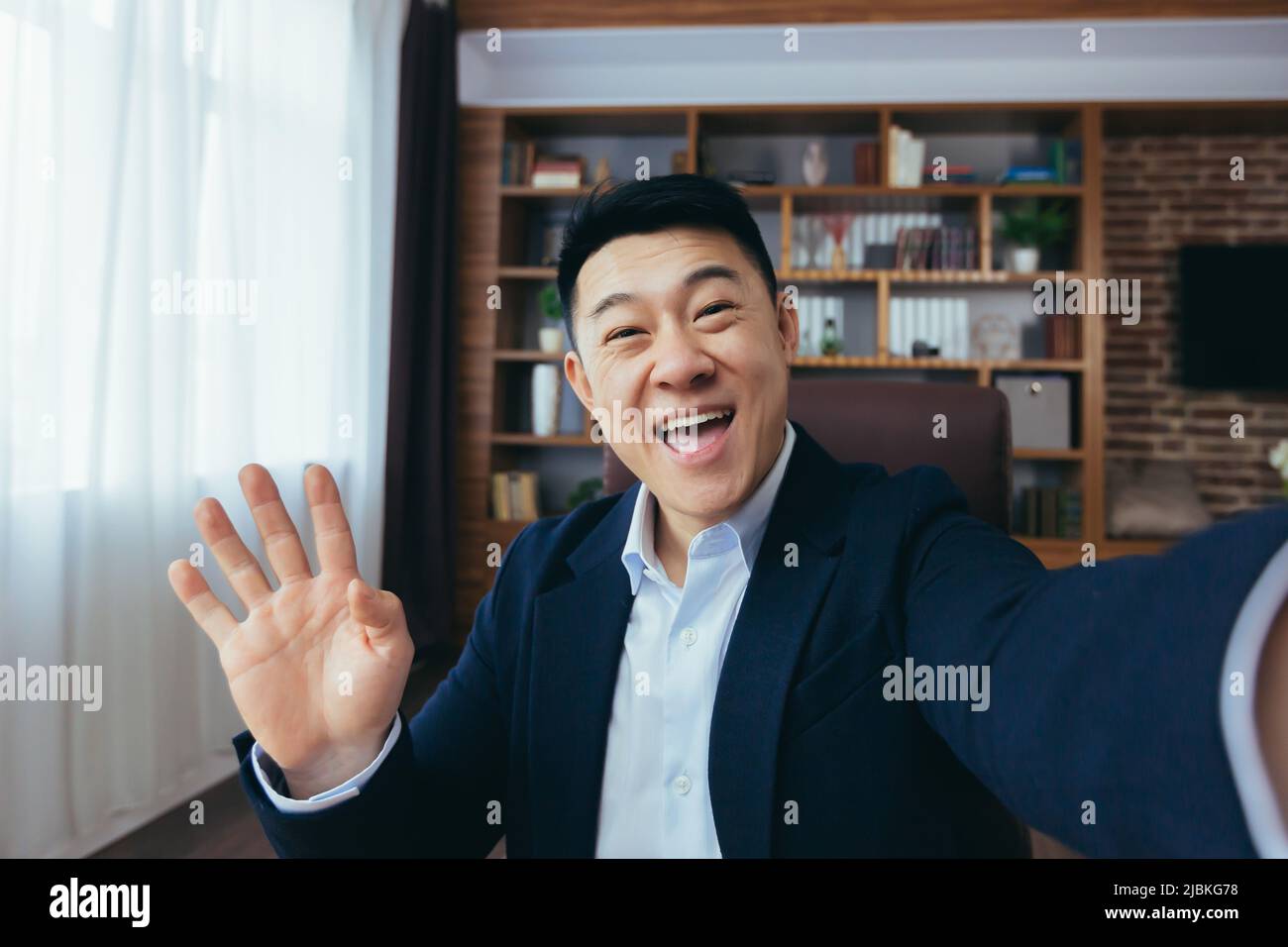 Cheerful and funny Asian businessman looks at the camera of a smartphone, talks on a video call, a man works in the office, waves a congratulatory ges Stock Photo
