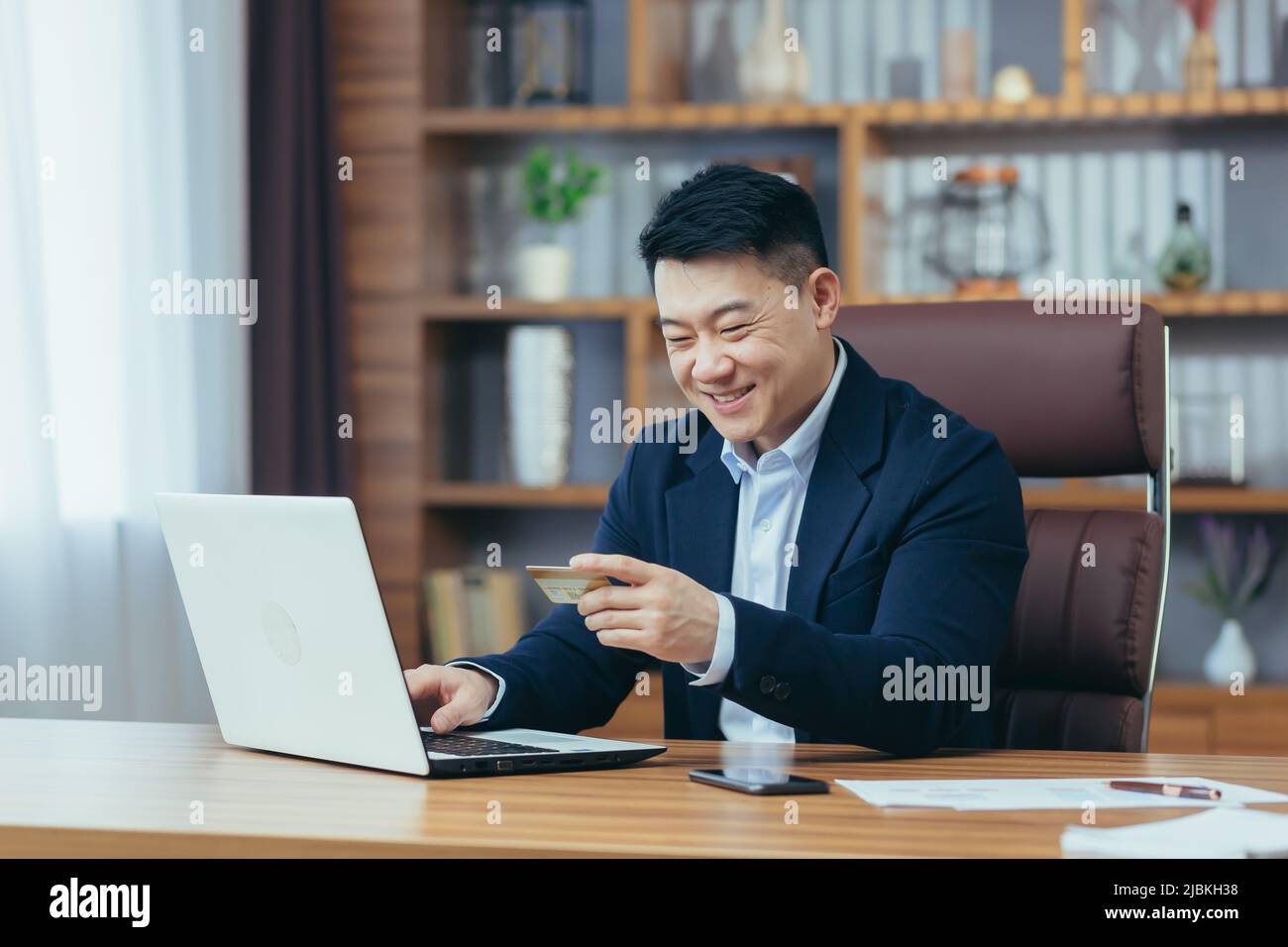 Successful Asian businessman carries out banking operations and transfers money from an electronic account, uses a laptop and a bank credit card Stock Photo