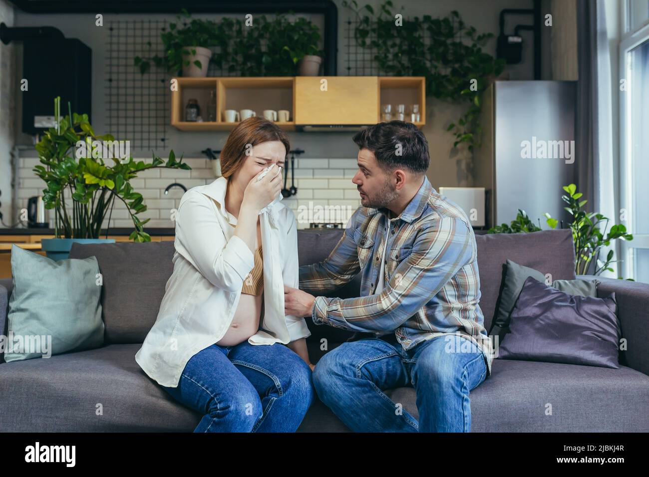 Young pregnant woman crying, wipes tears with a napkin. The man calms her down, supports her. Sitting at home on the couch. Emotional state of a pregn Stock Photo