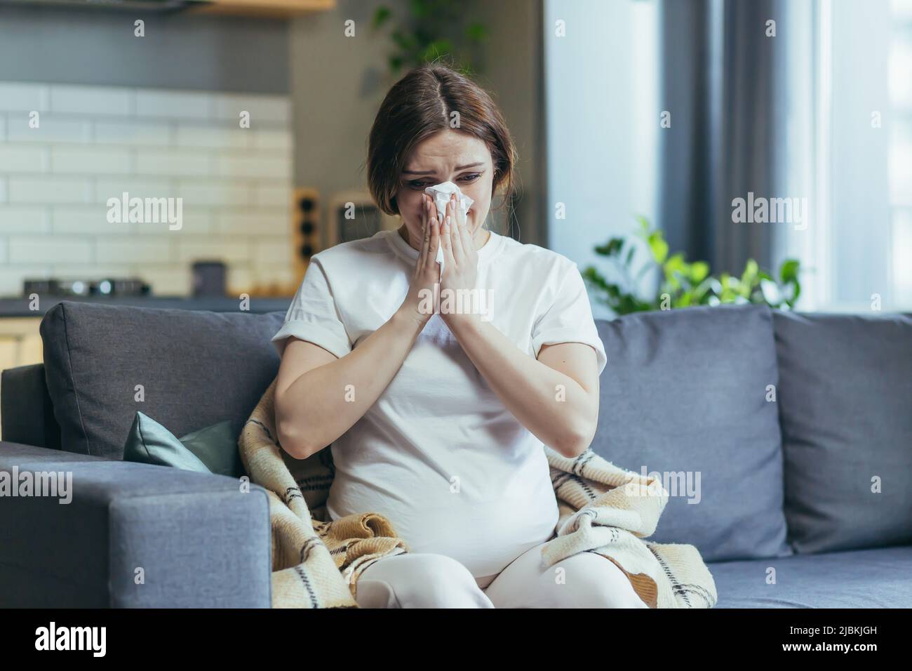 Sad pregnant woman at home sitting on the couch and crying, has a runny nose and allergies sneezes into a napkin Stock Photo