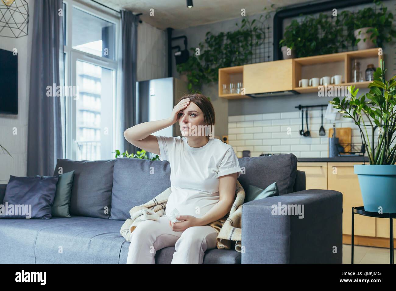 Feeling unwell. A pregnant woman is ill, has a headache, holds her hand behind her head. Sitting at home in home clothes on the couch Stock Photo