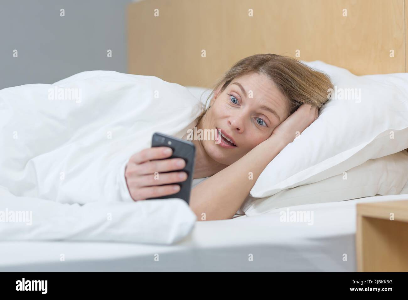 Woman in bed under the blanket shocked, reading good news from the