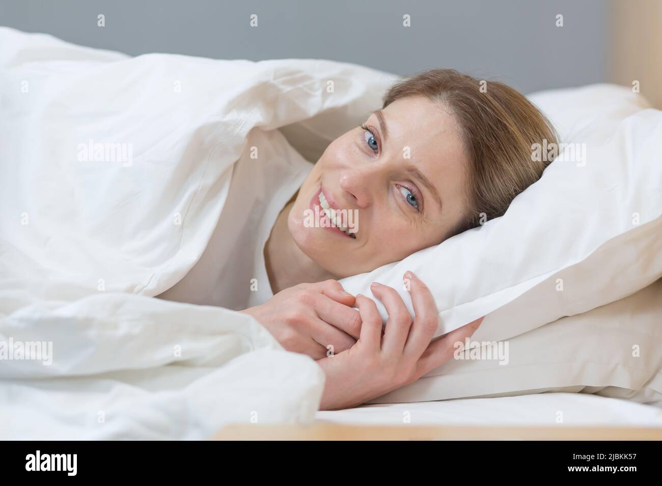 Close-up photo of portrait of woman sleeping in bed smiling and looking at camera in the morning Stock Photo