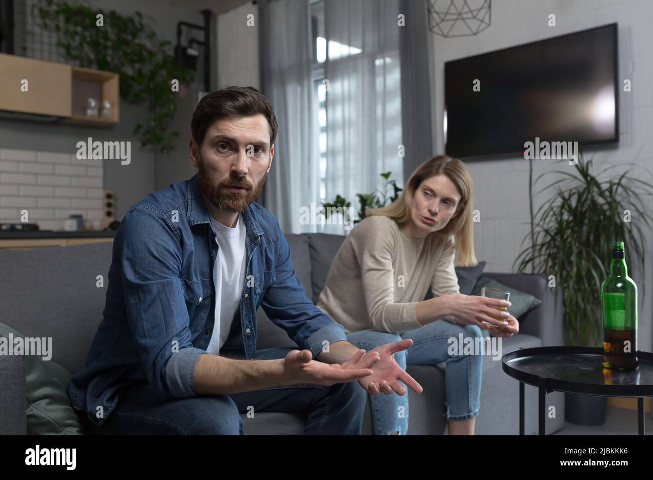 Female alcoholism, husband in despair, woman wife drinking hard alcohol, couple sitting on sofa in living room Stock Photo