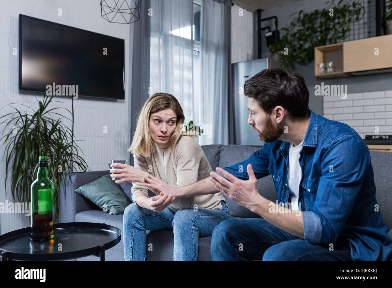Female alcoholism, family quarrel at home, wife drinking alcohol, husband trying to stop her, sitting on the couch Stock Photo