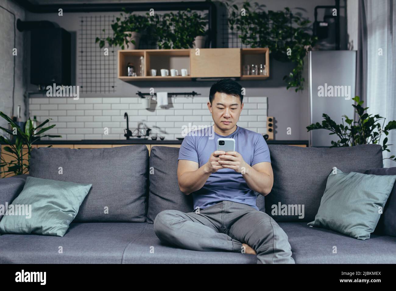 Concerned man reading news online looking at phone screen, Asian sitting on sofa at home, serious and sad Stock Photo