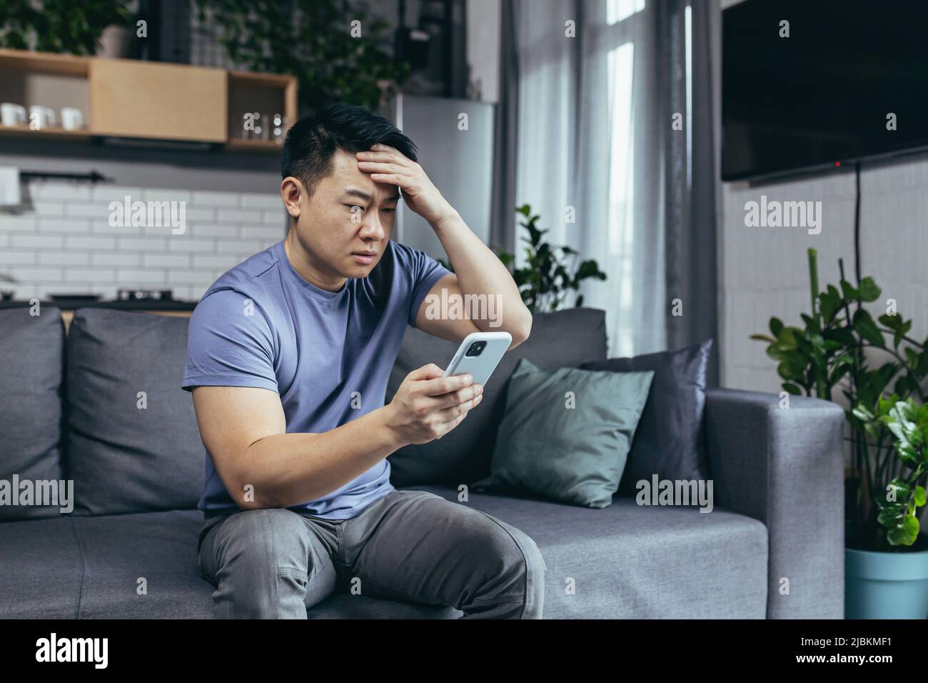 Concerned man reading news online looking at phone screen, Asian sitting on sofa at home, serious and sad Stock Photo