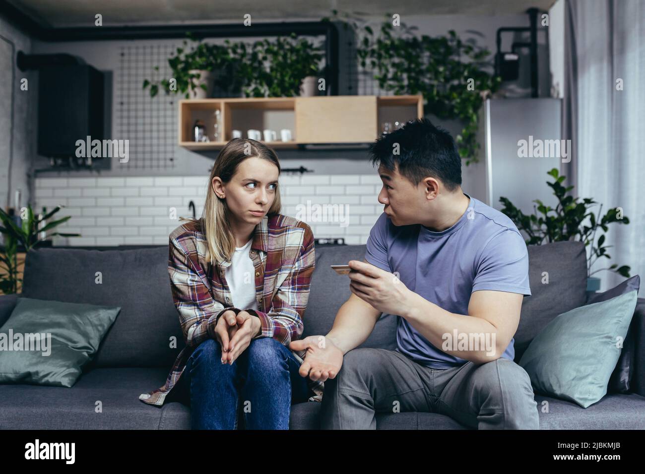 Asian man quarrels with his wife, accuses him of squandering family money, family quarrel, couple sitting on sofa, husband points to reduction of cred Stock Photo