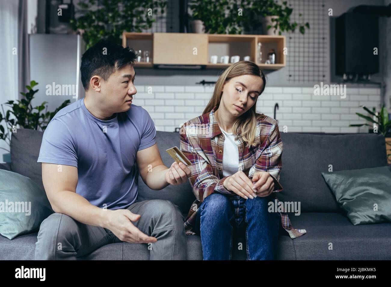 Asian man quarrels with his wife, accuses him of squandering family money, family quarrel, couple sitting on sofa, husband points to reduction of cred Stock Photo