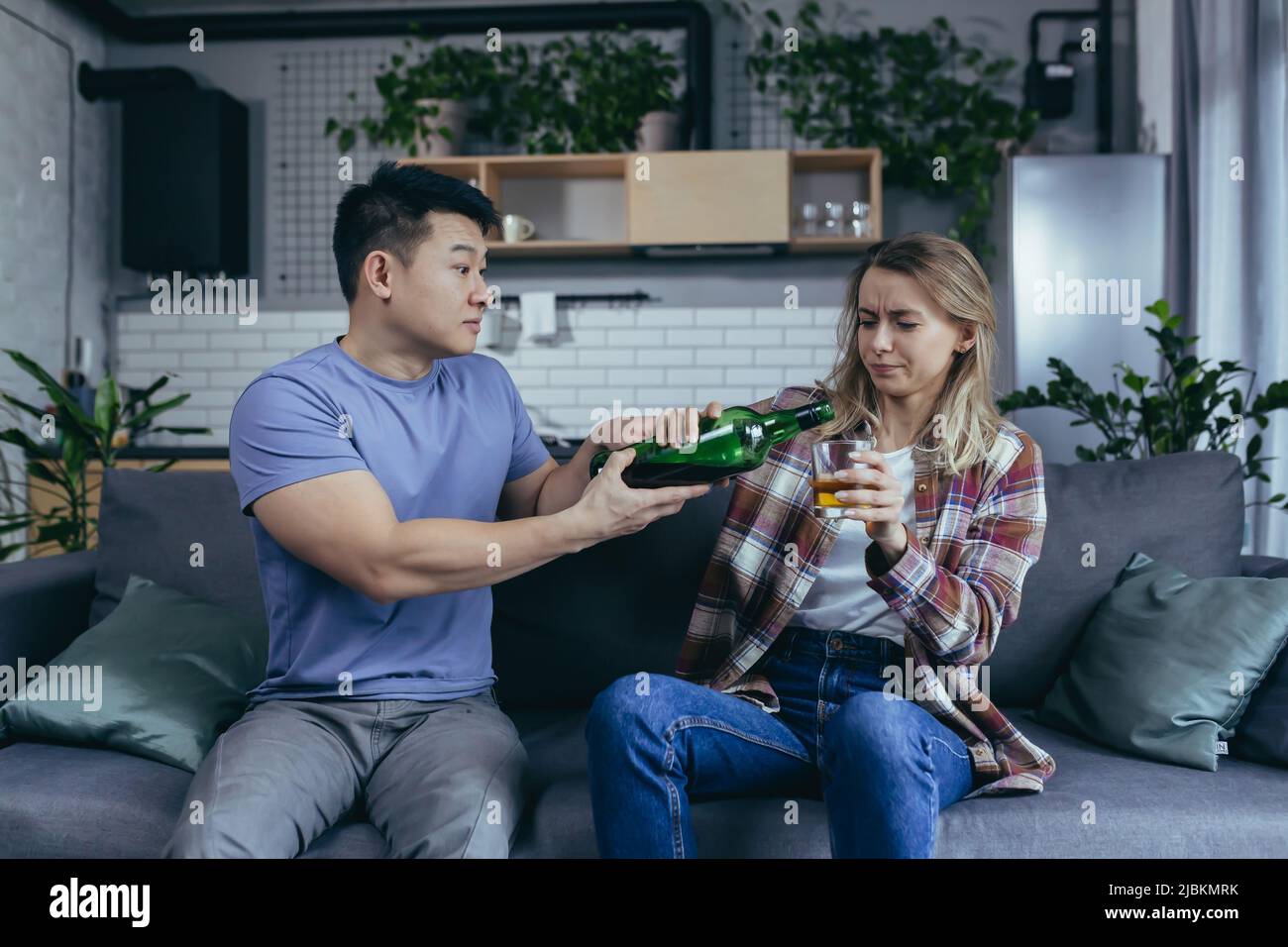 Alcoholic woman drinking hard alcohol at home, husband frustrated and annoyed, conflict in young family over alcohol Stock Photo