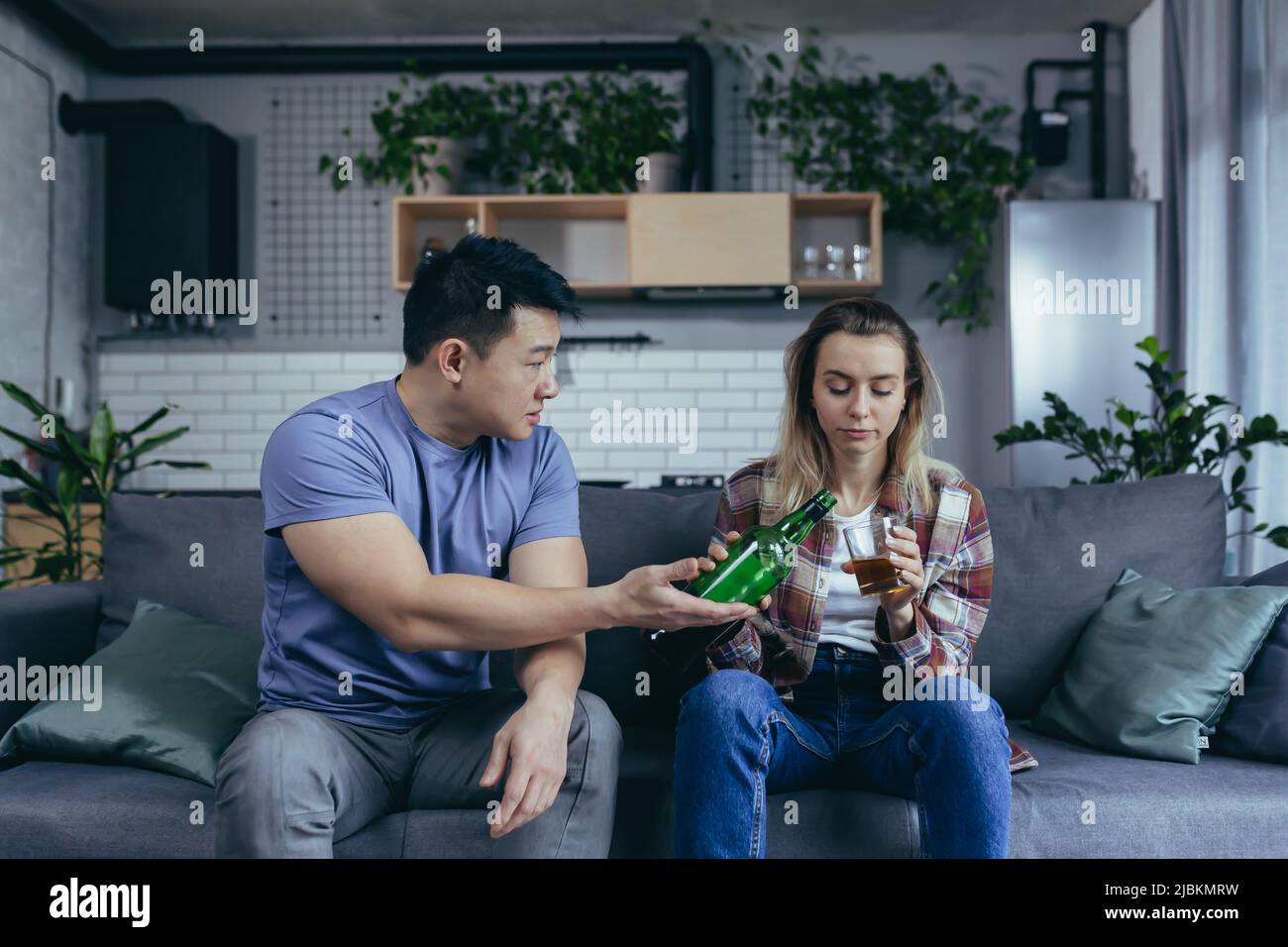 Alcoholic woman drinking hard alcohol at home, husband frustrated and annoyed, conflict in young family over alcohol Stock Photo