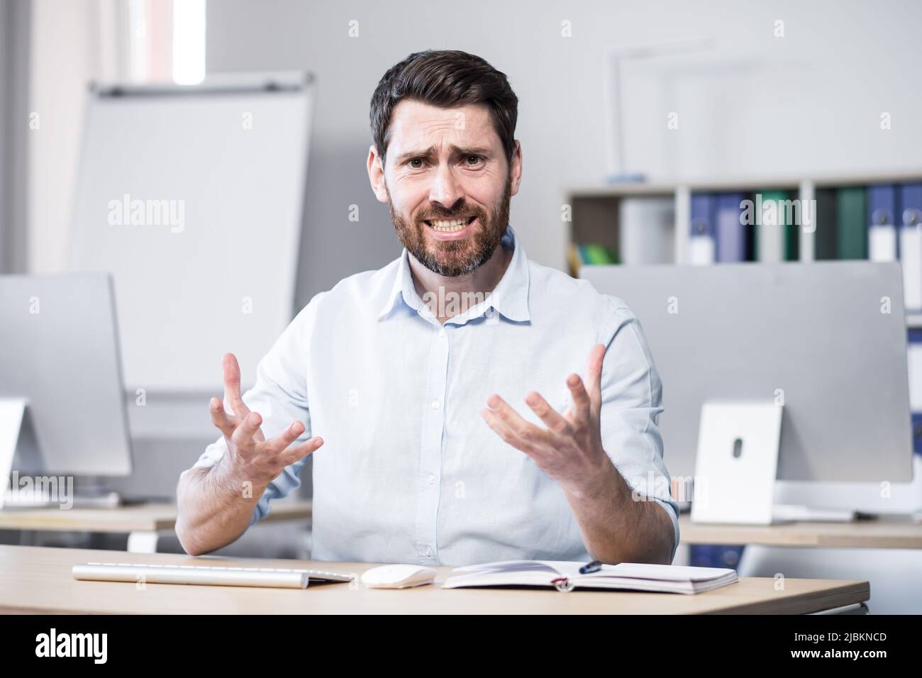 Young handsome successful business coach explains and tells on camera, gestures with his hands. Sitting at a computer desk in the office, looking at t Stock Photo