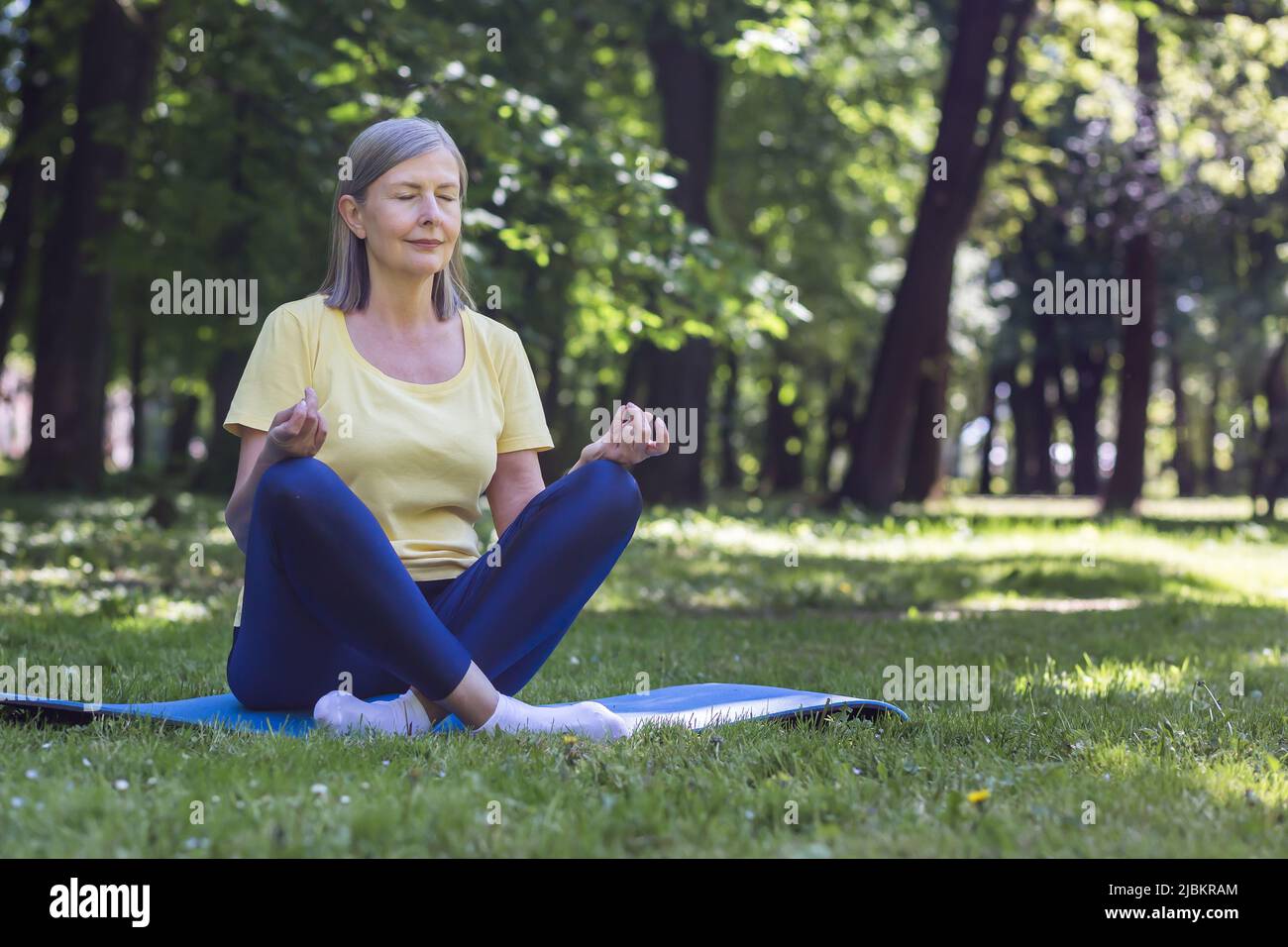 Senior gray-haired retired woman in the park meditates and performs breathing exercises, summer day in lotus position Stock Photo