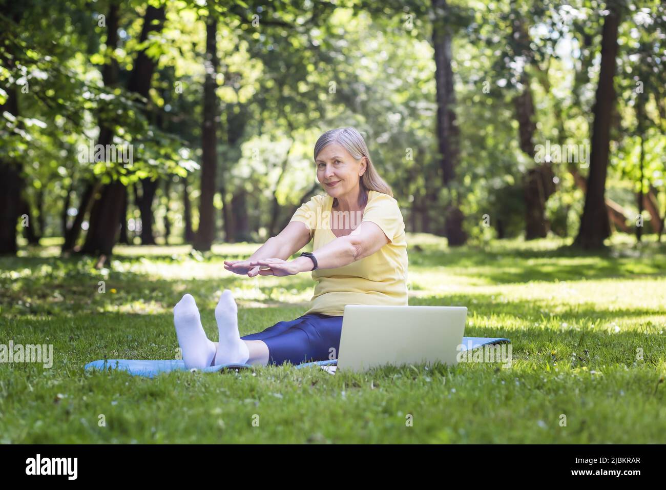 Active gray-haired woman in the park doing fitness sitting on the mat and grass, using a laptop for online classes in the park Stock Photo